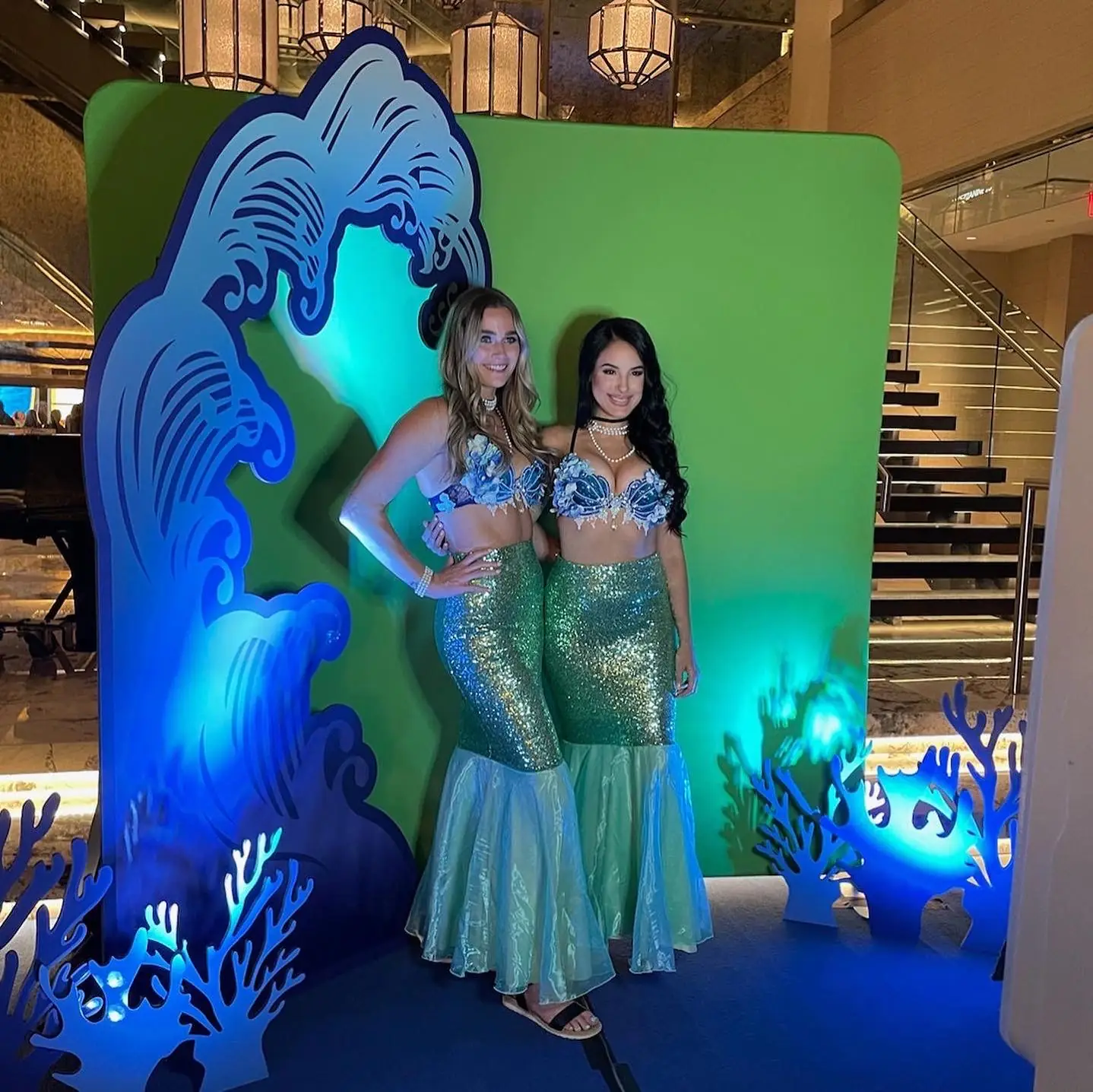 Two performers in mermaid costumes posing against the green screen backdrop at an under-the-sea themed hotel event